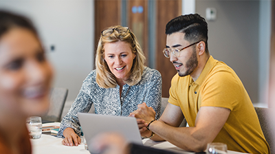 A man and a woman sitting at a table while looking at a laptop. A smiling woman out of focus in the forefront.