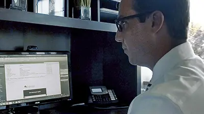 Man in glasses sitting at a desk looking at a computer in front of him.