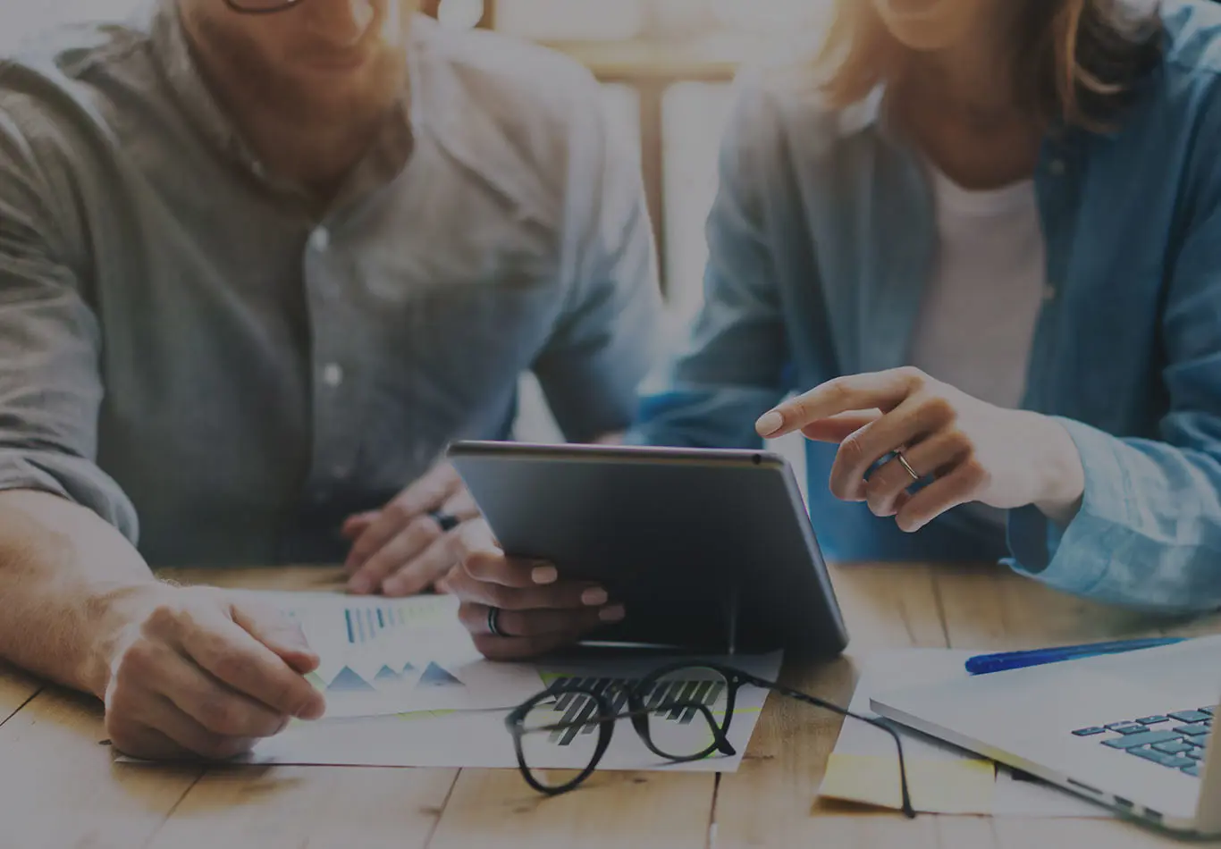 Two people are sitting at a table together. One person in a grey shirt has papers in front of them and the other person in a blue shirt is holding a tablet and has a computer in front of them