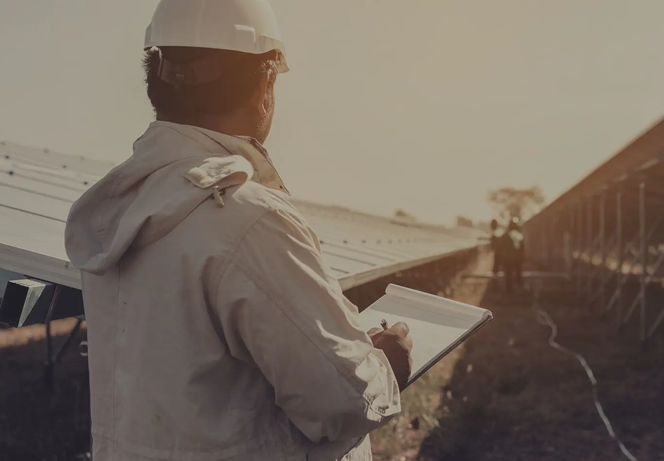 A person in a hard hat and jacket is holding a clip board and looking towards two other people and solar panels.