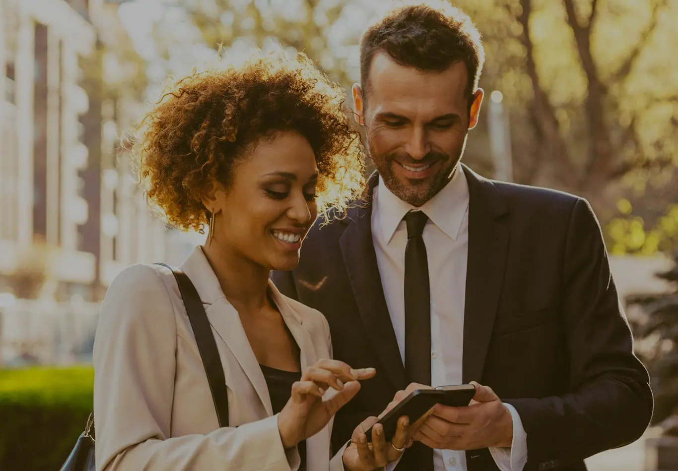 A business man and business woman are standing next to each other on the street. They are looking at their phones. 