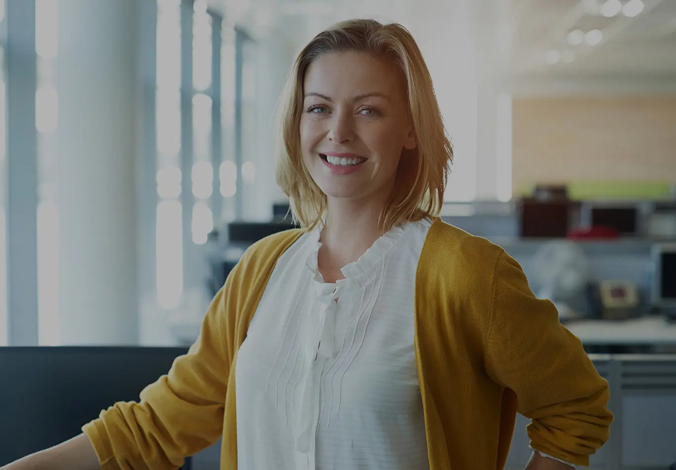 A blonde haired business woman in a white blouse and yellow cardigan is looking at the viewer. 