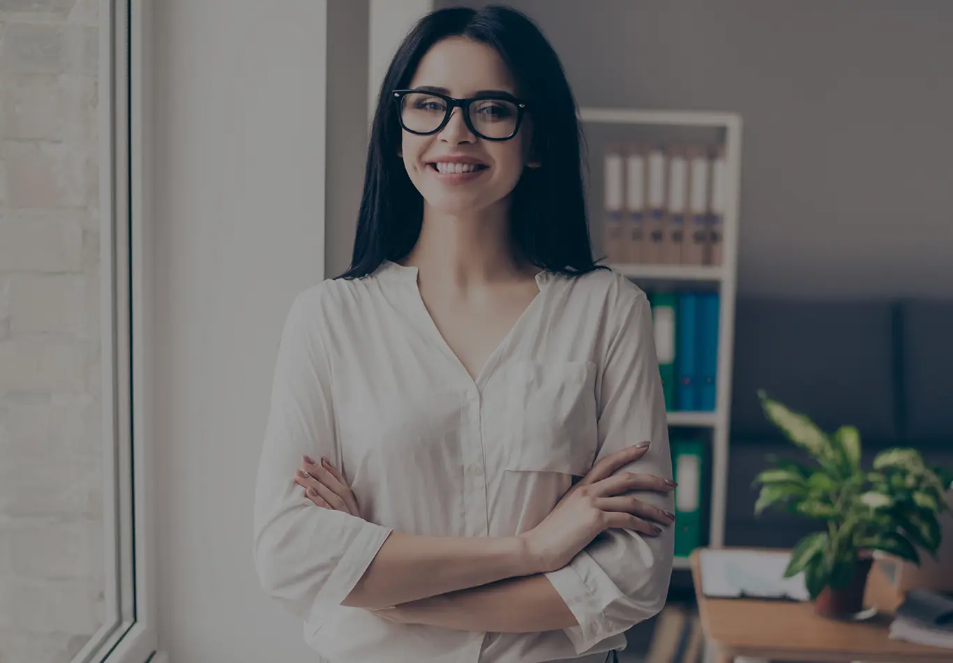 Woman in casual attire  in an office wearing large glasses with arms folded over her chest, she is smiling at the camera. 